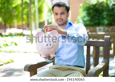 Closeup portrait, young business man putting coins in piggy bank, isolated outdoors trees background. Smart wise currency financial investment wealth decisions. Budget management and savings