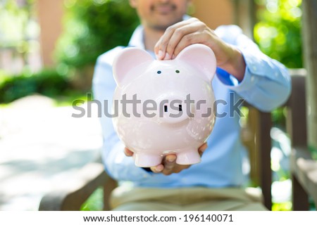 Closeup portrait, young business man putting coins in piggy bank, isolated outdoors trees background. Smart wise currency financial investment wealth decisions. Budget management and savings