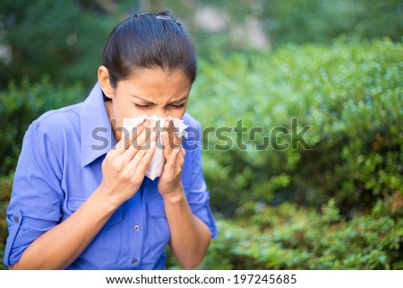 Closeup portrait, young woman in blue shirt with allergy or cold, blowing her nose with a tissue, looking miserable unwell very sick, isolated outside green trees background. Flu season, vaccination.