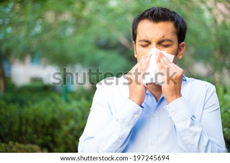 Closeup portrait of young man in blue shirt with allergy or cold, blowing his nose with a tissue, looking miserable unwell very sick, isolated outside green trees background. Flu season, vaccination.
