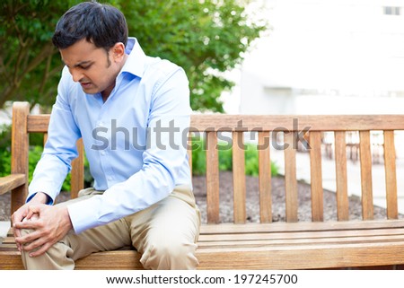 Closeup portrait, young handsome man in blue shirt, brown khakis, sitting on wooden bench in severe knee pain, isolated trees outside background. Negative human emotion