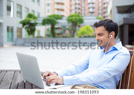 Closeup portrait, young handsome man in blue shirt typing away, listening to headphones, browsing digital computer laptop, isolated background of sunny outdoor, green trees, office background