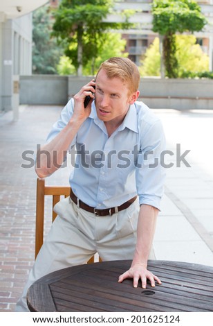 Closeup portrait, angry young man in blue shirt, annoyed by what he hears on cell phone, standing up with hand on table, isolated outdoors background