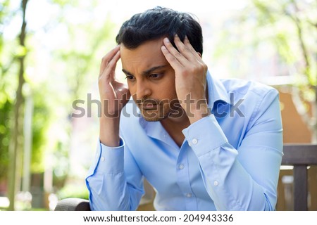 Closeup portrait, stressed young business man, hands on head with bad headache, isolated background of trees outside. Negative human emotion facial expression feelings.