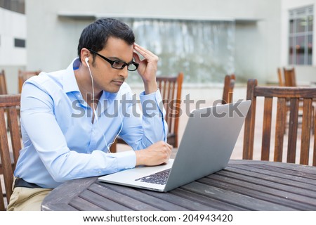 Closeup portrait, young man in blue shirt and black glasses, listening to headphones, browsing digital computer laptop, isolated background of sunny outdoor, gray building waterfall, with brown chairs