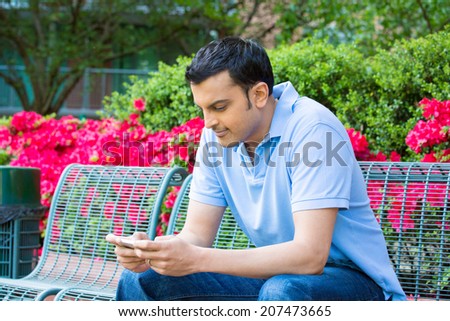 Closeup portrait, young happy man in blue shirt sitting on green bench, engrossed, checking his cellphone, isolated on background of red flowers outside