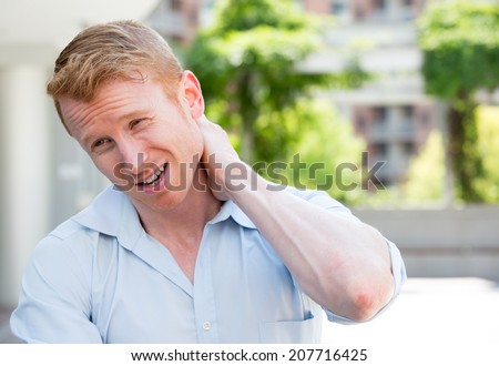 Closeup portrait, young man in blue shirt with spinal neck pain in thoracic vertebrae after long hours of work and studying, isolated outside outdoors background.  Lack of ergonomic support