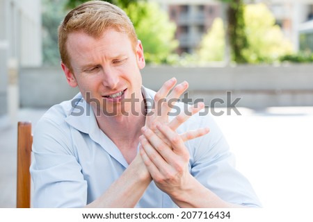 Closeup portrait, young man having acute bad joint pain in his hands, writer\'s cramp, massaging them, sitting in chair, isolated outdoors background