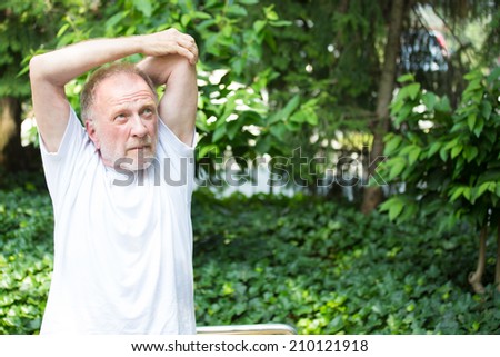 Closeup portrait, senior mature man in white shirt, stretching arms, isolated green trees background. Warming up