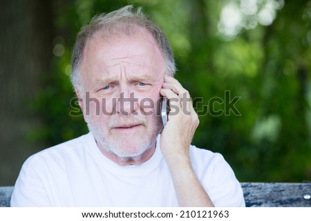 Closeup portrait, senior mature man in white t-shirt sitting on bench, having serious conversation on cell phone, isolated outside outdoors background