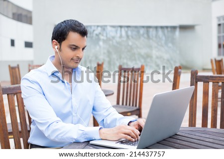 Closeup portrait, young handsome man in blue shirt typing away, listening to headphones, browsing digital computer laptop, isolated background of sunny outdoor, brown chairs background