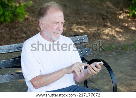 Closeup portrait of bold elderly man in white shirt, squinting, checking smartphone, sending text message, seated on a bench, isolated outdoor background.