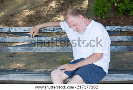 Closeup portrait bald old man in white shirt, blue shorts, exhausted, resting on a bench, looking down, trying to take a power nap, isolated outdoor outside background