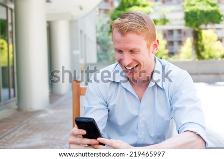 Closeup portrait, young happy businessman in blue shirt sitting, checking his cellphone, isolated on background of a city building, trees, on a sunny autumn day. Corporate life success.