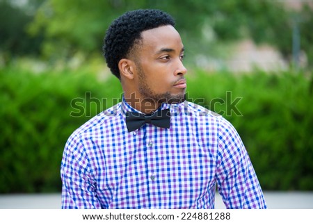 Closeup side profile portrait, handsome business man in blue shirt, bow tie, relaxing outside in a park during sunny day, looking away, isolated outdoors green tress background.