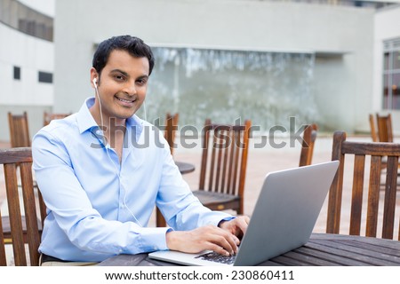 Closeup portrait, happy young handsome man in blue shirt typing away, browsing digital computer laptop, isolated background of sunny outdoor, waterfall, brown chairs, office background