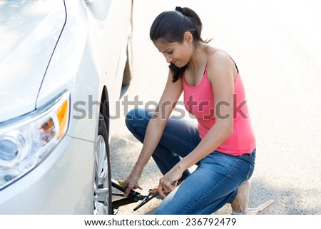 Closeup portrait, young woman in pink tanktop and blue jeans fixing flat tire with jack and tire iron, isolated green trees and road outside background. Roadside assistance concept