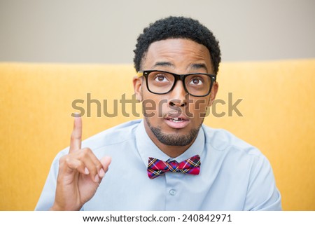 Closeup portrait young man with big black glasses blue shirt and bow tie has an idea, pointing with finger up looking up, isolated  yellow gray background.