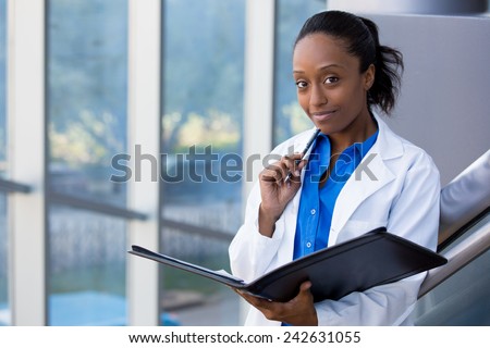 Closeup headshot portrait of friendly, smiling confident female doctor, healthcare professional with labcoat, holding pen to face and holding notebook pad. Isolated hospital clinic background.