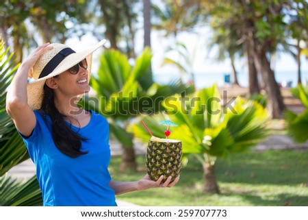 Closeup portrait, woman in blue shirt and brown straw hat holding pineapple mixed drink with rum at resort, isolated background of green palm trees