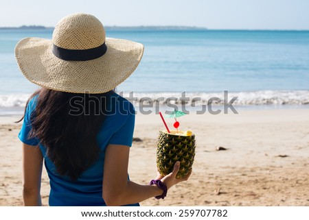 Closeup portrait, woman in blue shirt and brown hat holding pina colada rum pineapple mixed drink with straw and tiny umbrella, while looking out towards beach and ocean