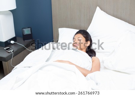 Closeup portrait, young woman sound asleep, catching Zs in bed, white pillows, charging cell phone and with alarm clock and lamp shade. Tackling everyday stress and anxiety with cat nap
