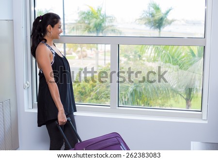 Closeup portrait, young woman in black shirt holding maroon suitcase, packed and ready to go, looking out glass window with palm trees background