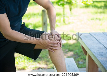 Closeup cropped portrait, man in black shirt and shorts holding knee in severe pain, isolated trees and picnic bench outside background.