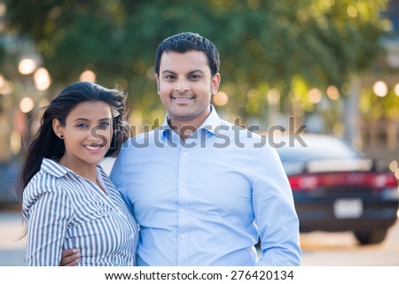Closeup portrait, attractive wealthy successful couple in blue shirt and striped outfit holding each other smiling, isolated outside green trees and black car background.