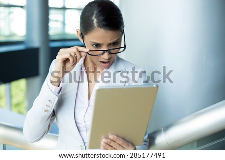Closeup portrait, upset young woman, astonished surprised, wide open mouth, large eyes in black glasses by what she sees on her gray silver tablet pad, isolated indoors office background.