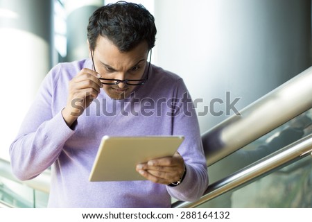 Closeup portrait, dumbfounded, flabbergasted man in black eyeglasses and purple sweater, squinting eyes, raising eyebrows, looking closely at tablet, isolated indoors office background