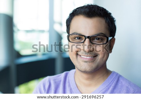 Closeup headshot portrait, smiling happy handsome man in purple sweater v-neck, wearing black glasses, isolated inside office background.