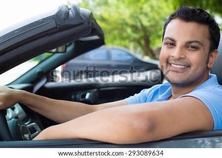 Closeup portrait, happy young smiling handsome man in blue polo shirt in his new black sports car, relaxing, looking at camera, isolated on outdoors background with vehicle and green trees.