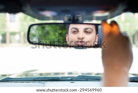 Closeup portrait, young handsome egotistical man in blue polo shirt looking at mirror reflection showing kisses and duck face, admiring his appearance inside interior black sports car
