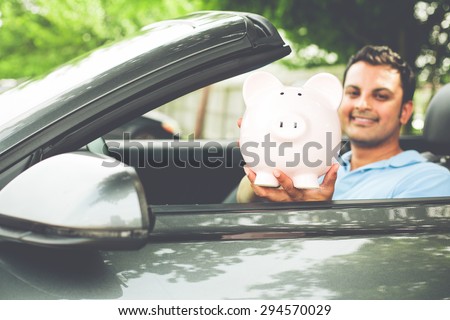 Closeup portrait, handsome young man in blue polo shirt showing pink piggy bank inside new sports car, isolated outdoors background.  Happy to have good low apr interest rates, finance rebate deals