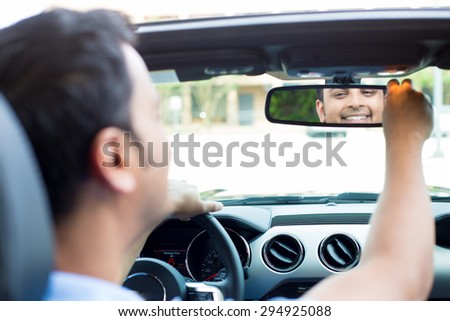 Closeup portrait, happy young man driver looking at rear view mirror smiling, adjusting image reflection, isolated interior car windshield background. Positive human expression body language