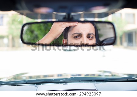 Closeup portrait, funny young man driver looking at rear view mirror looking at hair loss issues widow\'s peak or worried, isolated interior car windshield background