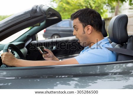 Closeup portrait, funny young shocked man wide open mouth, texting on cellphone while driving in gray black charcoal sports car, isolated outside background.