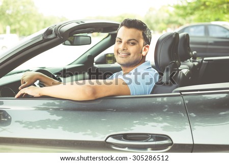 Closeup portrait, happy young smiling man in blue polo shirt in his new black sports car, relaxing, looking at camera, isolated on outdoors background with vehicle and green trees. Retro vintage look