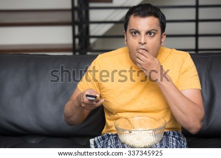 Closeup portrait, young man in yellow t-shirt, sitting on black leather couch, watching TV, holding remote, surprised at what he sees, munching popcorn, isolated indoors flat background