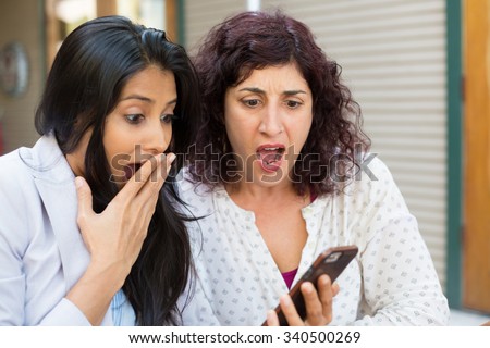Closeup portrait two surprised girls looking at cell phone discussing latest gossip news, shopping, flabbergasted at what they see, isolated outdoors background