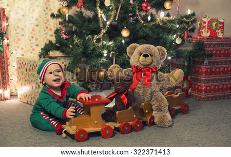 Baby in elf costume playing with old wooden train and soft toy bears under the Christmas tree, vintage.