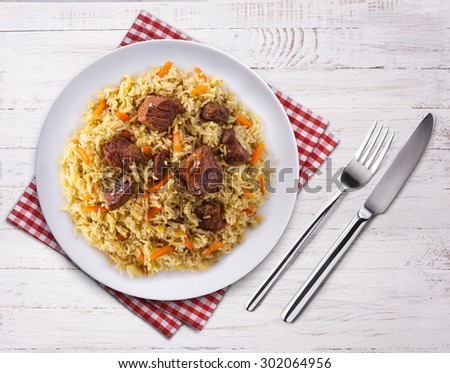Fragrant pilaf with meat and vegetables close up on a plate. Top view on wooden table. Fork, knife and towel.