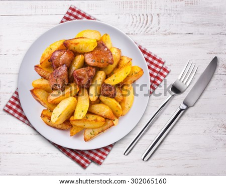 Spicy fried potatoes with herbs and spices served in white plate. Top view on wooden table. Fork, knife and towel.