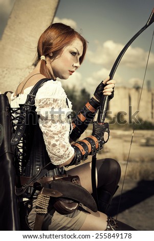 Attractive severe redhair woman warrior holding in her hands bow and arrow on the industrial background.
