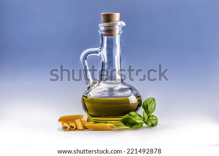 Glass bottle carafe with olive oil penne pasta and basil leaves on white and blue background.