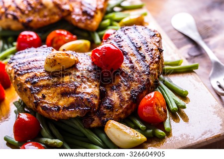 Grilled chicken breast in different variations with cherry tomatoes, green French beans, garlic, herbs, cut lemon on a wooden board or teflon pan. Traditional cuisine. Grill kitchen.