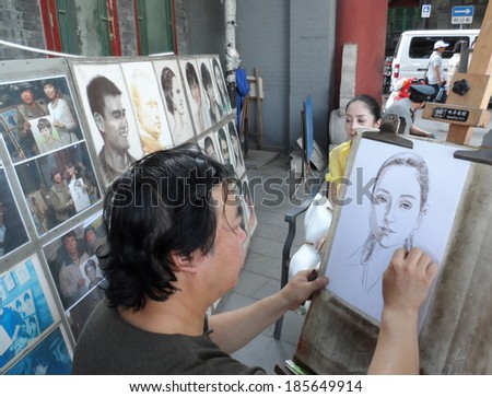 Beijing, China Ã¢Â?Â? July 5, 2013: Street artist draw a portrait of woman in a park of Beijing, China. The woman sits on a chair. The artist shows some of his previous work on the stand.
