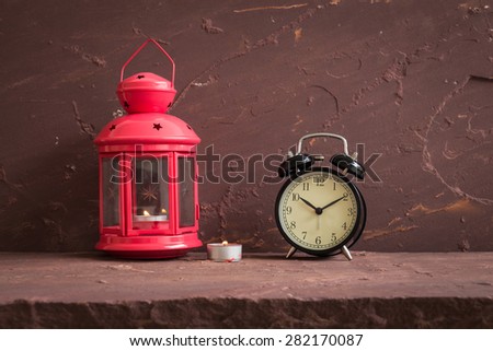 Red lantern, candle and black vintage clock on brown stone table over stone grunge background.