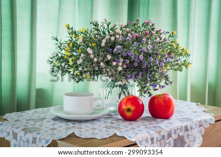 Bouquet of flowers with cup of coffee and red apple on a wooden table over green curtain background in the bedroom